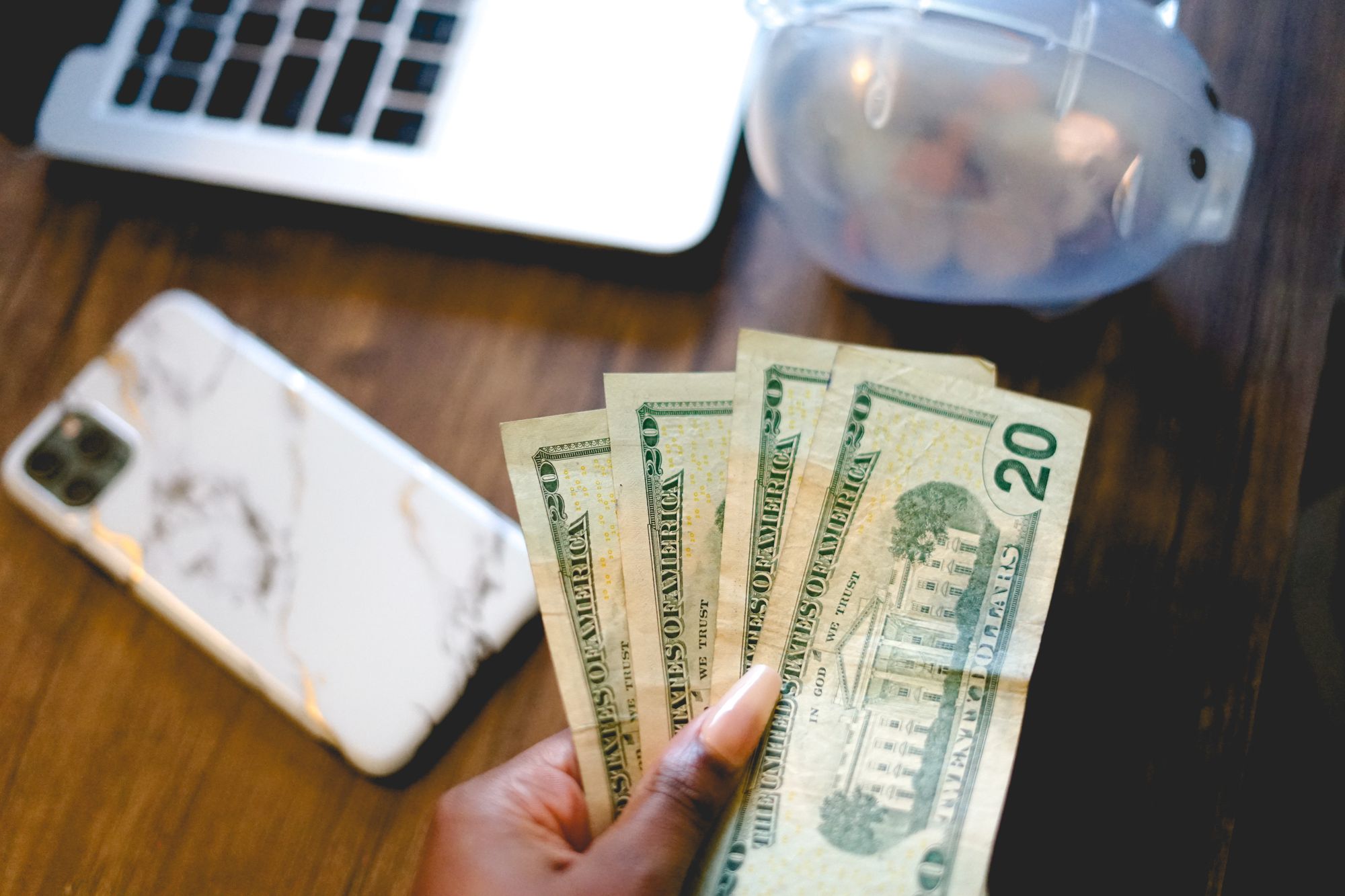 A woman off-screen holding $20 bills, with a piggy bank and phone on her desk.